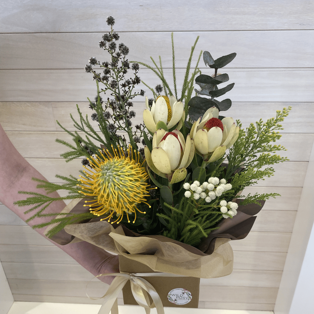 Bouquet of flowers from Wild Posy with a yellow protea held by a hand against a light wooden panel background